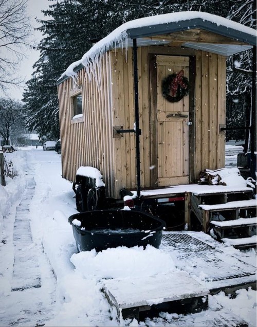 A cozy wooden cabin on wheels surrounded by snow, with a wreath on the door and a black tub nearby.