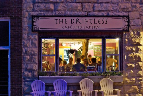 A cozy café and bakery named "The Driftless," with people dining inside, viewed through a warmly lit window.