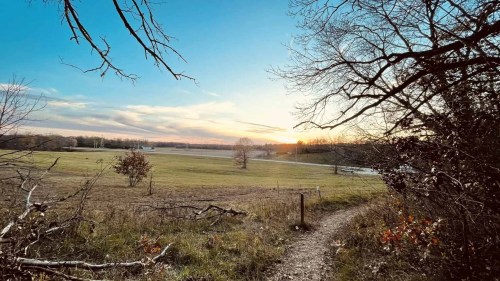A serene landscape at sunset, featuring a grassy field, trees, and a winding path under a colorful sky.