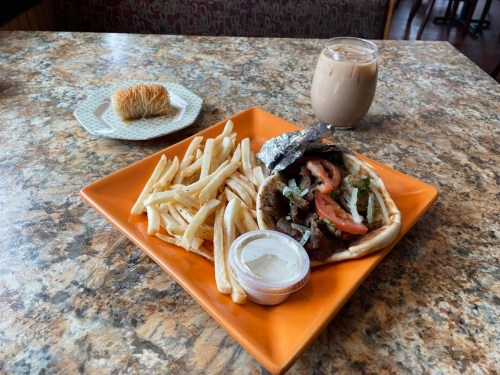 A plate with a sandwich, fries, and a dipping sauce, alongside a glass of iced coffee and a pastry on a table.