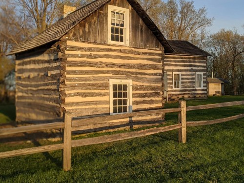 A rustic log cabin with a wooden fence, surrounded by grass and trees, illuminated by warm evening light.