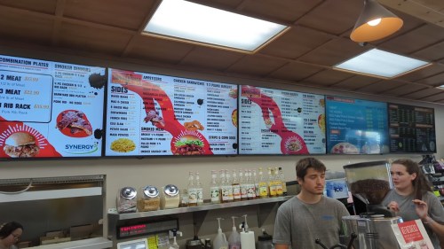 Menu boards displaying food options and prices at a restaurant, with staff members in the foreground.