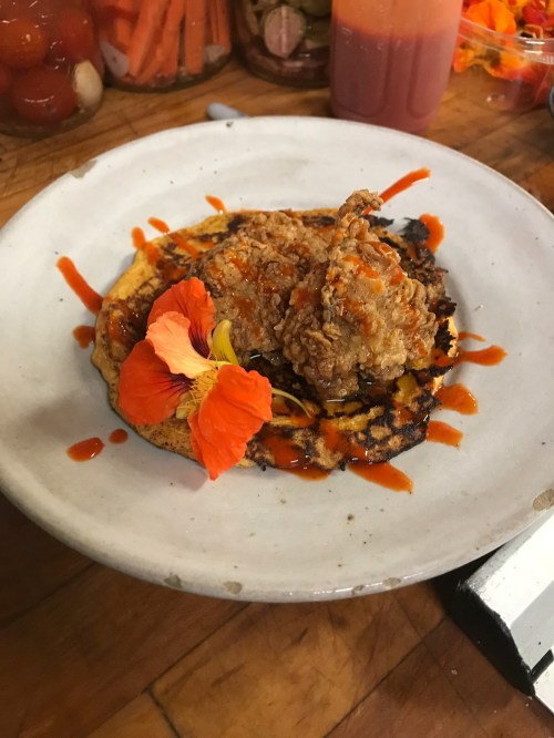 A plate with a crispy fried chicken piece on a corn cake, drizzled with sauce and garnished with an edible flower.