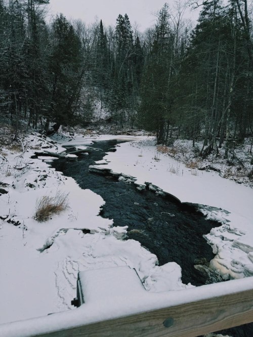 A serene winter scene of a river winding through a snowy forest, with ice patches on the water's surface.