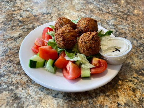 A plate of falafel balls on a bed of lettuce, tomatoes, and cucumbers, served with a side of dipping sauce.