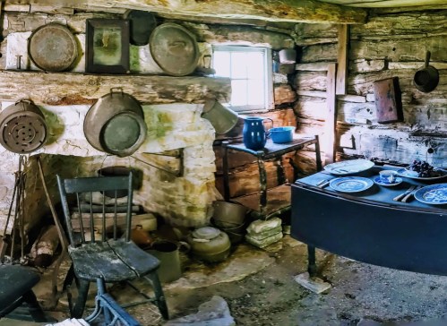A rustic kitchen with a wooden table, old cookware, a fireplace, and a chair, showcasing a cozy, historical atmosphere.