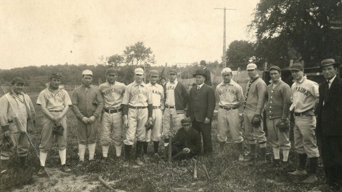 A vintage baseball team poses for a photo, wearing uniforms and hats, with a few men in suits standing nearby.