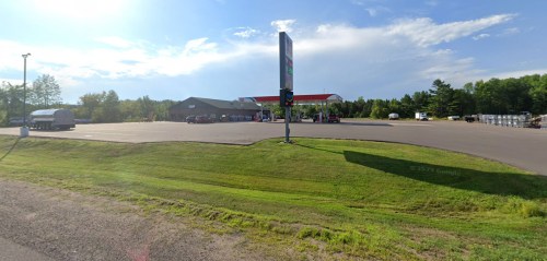 A gas station with a large sign, surrounded by green grass and trees under a partly cloudy sky.