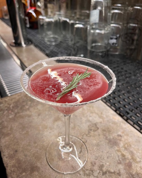 A cocktail in a glass with a salted rim, garnished with a sprig of rosemary, on a bar counter.