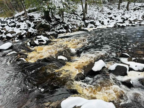 A snowy riverbank with flowing water and rocks, surrounded by trees in a winter landscape.