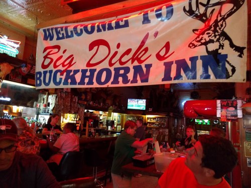 A colorful banner reading "Welcome to Big Dick's Buckhorn Inn" hangs in a lively bar with patrons and a TV in the background.