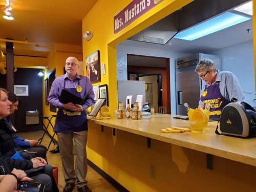 Two men in purple shirts and aprons stand behind a counter in a mustard-themed restaurant, with customers seated in front.