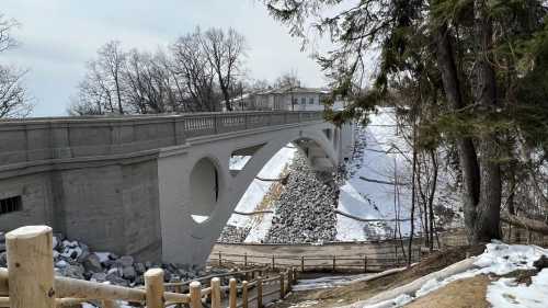 A curved concrete bridge spans a snowy landscape, surrounded by trees and rocky terrain.