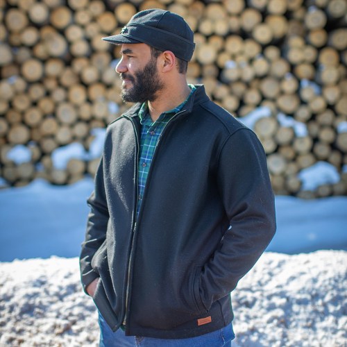 A man in a black jacket and cap stands outdoors, with a background of stacked logs and snow.