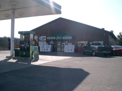 A gas station with a wooden building, featuring an ice cream shop and espresso stand, surrounded by parked vehicles.