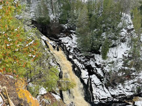 A snowy landscape featuring a waterfall cascading down rocky cliffs, surrounded by evergreen trees.
