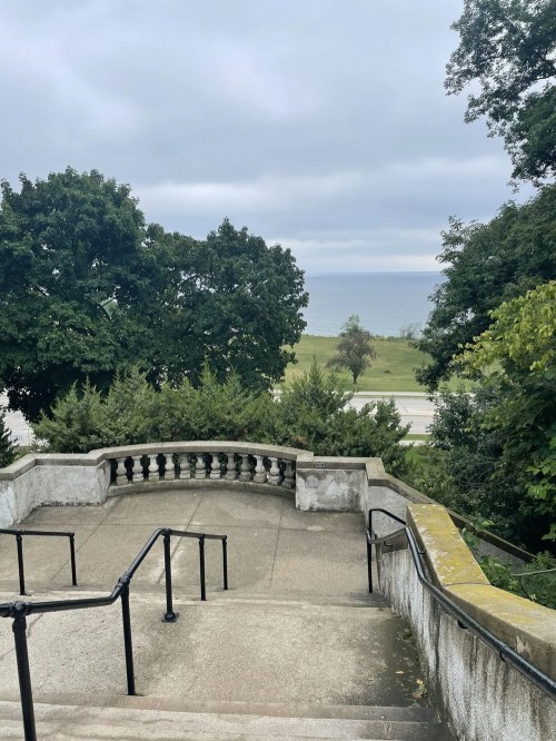 A view from stone steps leading down to a grassy area and a lake, framed by trees under a cloudy sky.