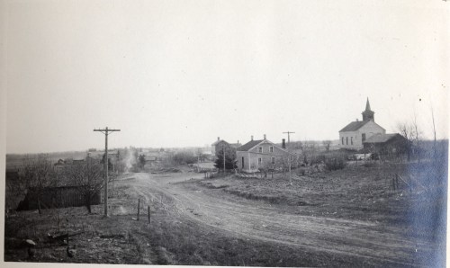 A rural landscape featuring a dirt road, a few houses, and a church in the distance, with sparse trees and fields.