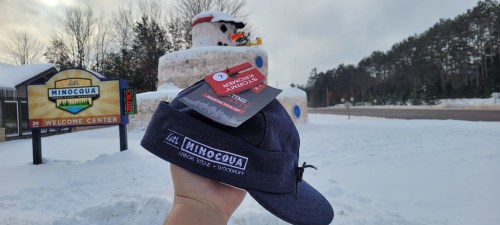 A person holds a navy blue visor in front of a snowman and a welcome sign for Minocqua, Wisconsin, in winter.