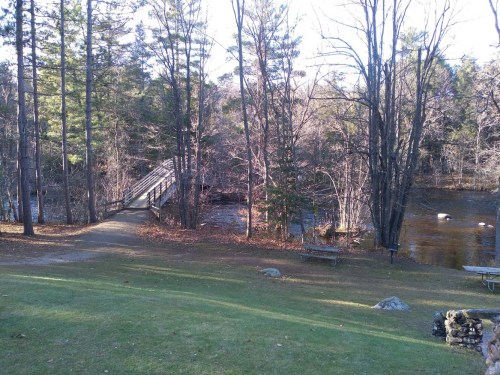 A serene landscape featuring a wooden bridge over a river, surrounded by trees and a grassy area.