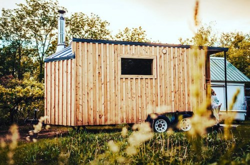 A wooden tiny house on wheels, surrounded by greenery, with a chimney and a large window.