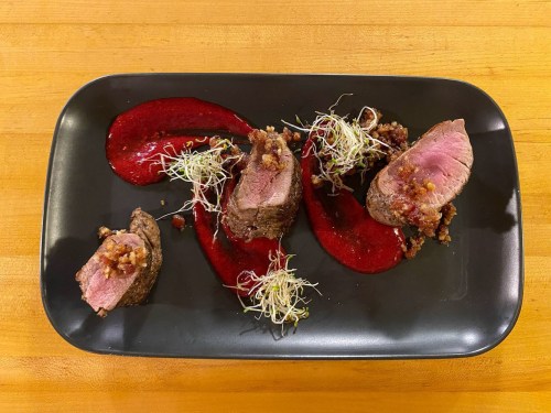 A black plate with sliced beef, garnished with sprouts and drizzled with a red sauce.