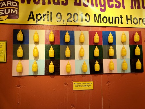 A colorful display of yellow mustard bottles arranged on a wall, with a sign warning not to squeeze or touch them.