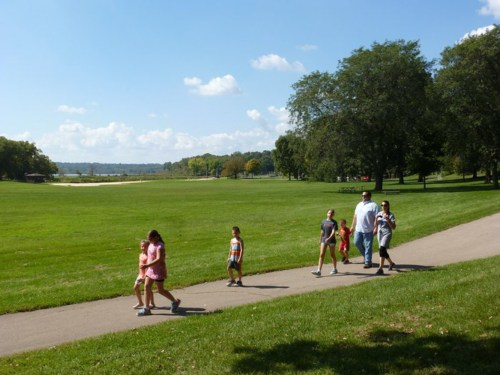 A group of children and adults walking along a path in a sunny park with green grass and trees in the background.
