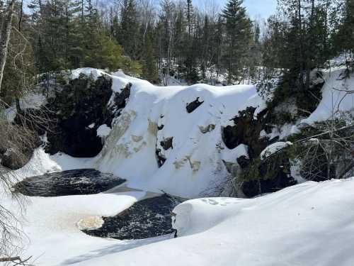 A snowy landscape featuring a waterfall cascading into a dark pool, surrounded by trees and rocky cliffs.