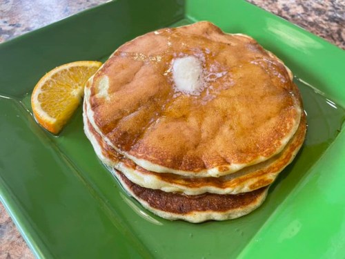 Three fluffy pancakes stacked on a green plate, topped with a pat of butter and drizzled with syrup, alongside a slice of orange.
