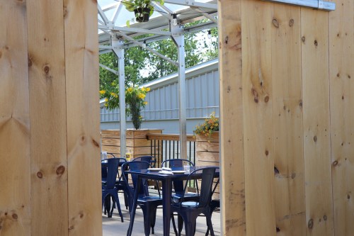 View through wooden panels to a patio with tables and chairs, surrounded by greenery and flowers.