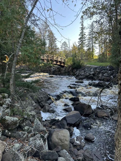 A serene river flows over rocks, with a wooden bridge in the background surrounded by trees and a clear blue sky.