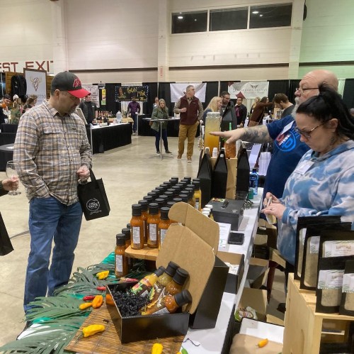 A vendor showcases products to a customer at a market event, with various bottles and displays in the background.