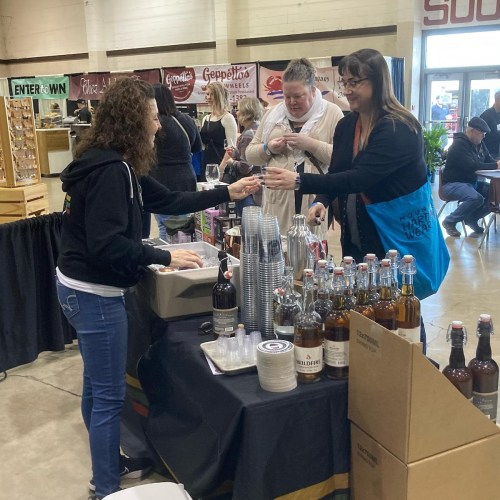 A vendor hands a drink to a customer at a market, with bottles and cups displayed on the table.