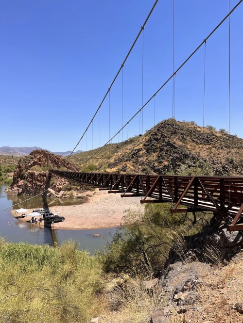 A long suspension bridge spans a river, surrounded by rocky hills and clear blue skies.