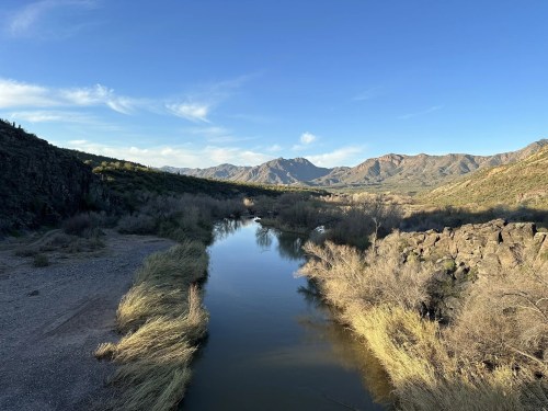 A serene river flows through a mountainous landscape, surrounded by dry vegetation and clear blue skies.