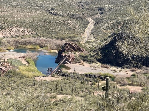 A scenic view of a river surrounded by rocky hills and desert vegetation, featuring a wooden bridge in the foreground.