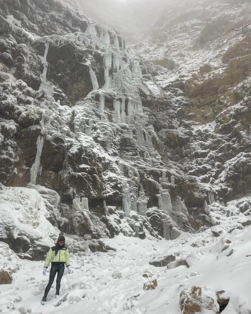 A person stands in front of a snowy, icy cliff with frozen waterfalls cascading down the rocky surface.