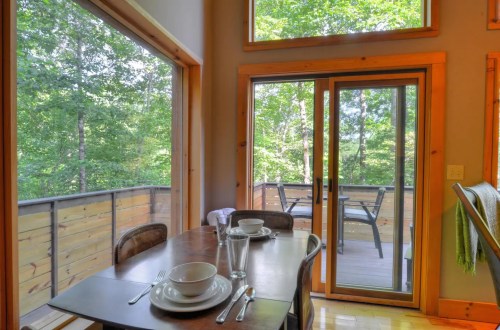 A cozy dining area with a wooden table set for two, overlooking a deck and lush green trees outside.