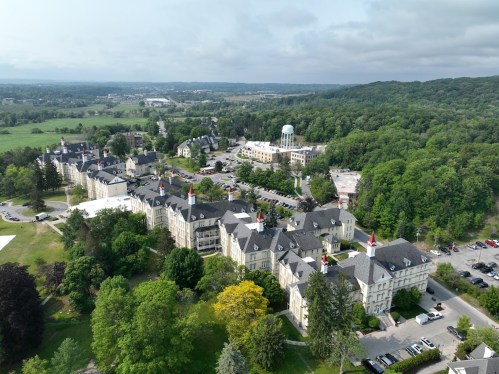 Aerial view of a historic building complex surrounded by greenery and hills, with a distant view of a water tower.