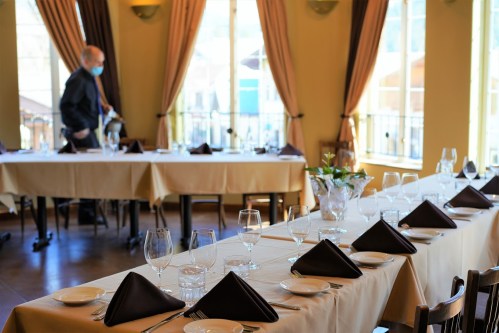A dining room set for a meal, featuring tables with white tablecloths, glassware, and a masked server in the background.
