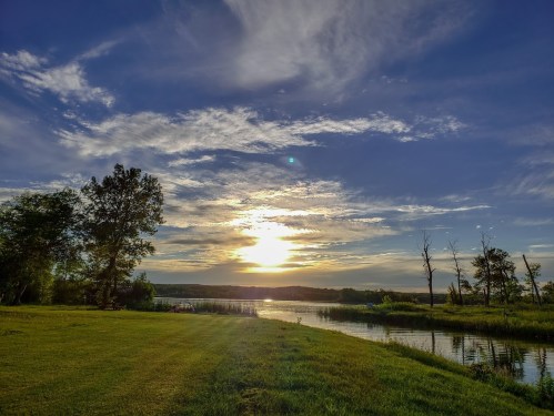A serene landscape featuring a sunset over a river, with lush green grass and trees under a partly cloudy sky.