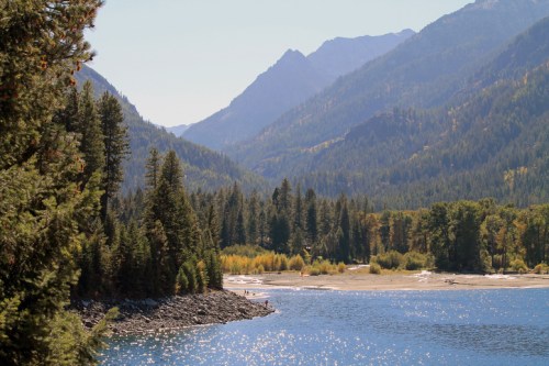 A serene landscape featuring mountains, a calm lake, and a sandy beach surrounded by lush trees.