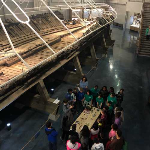 A group of people gathers around a table in a museum, with a large ship model displayed in the background.