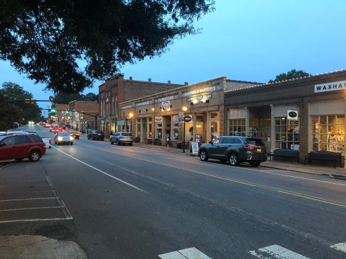 A quiet street lined with shops and cars, illuminated by warm lights at dusk. Trees frame the scene.