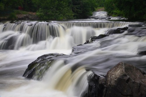 A serene waterfall cascades over rocky ledges, surrounded by lush greenery and flowing water.