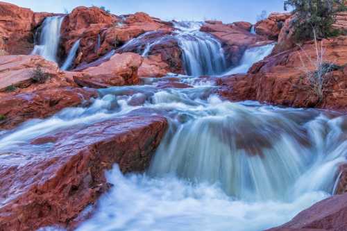 A serene waterfall cascades over red rock formations, surrounded by greenery and a soft blue sky.