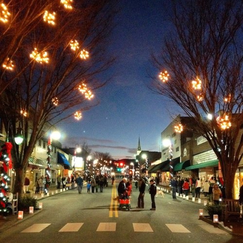 A festive street scene at dusk, with holiday lights, people walking, and shops decorated for the season.