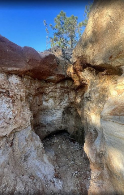 A rocky cave entrance surrounded by earthy tones and trees under a clear blue sky.