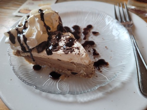 A slice of creamy dessert topped with whipped cream and chocolate drizzle on a glass plate. Fork beside it.